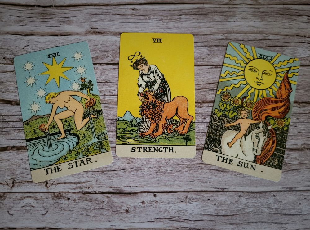 Photo of tarot cards. Depicted are The Star, The Strength and The Sun card. All three lying in a slight upwards curve on a wooden background
